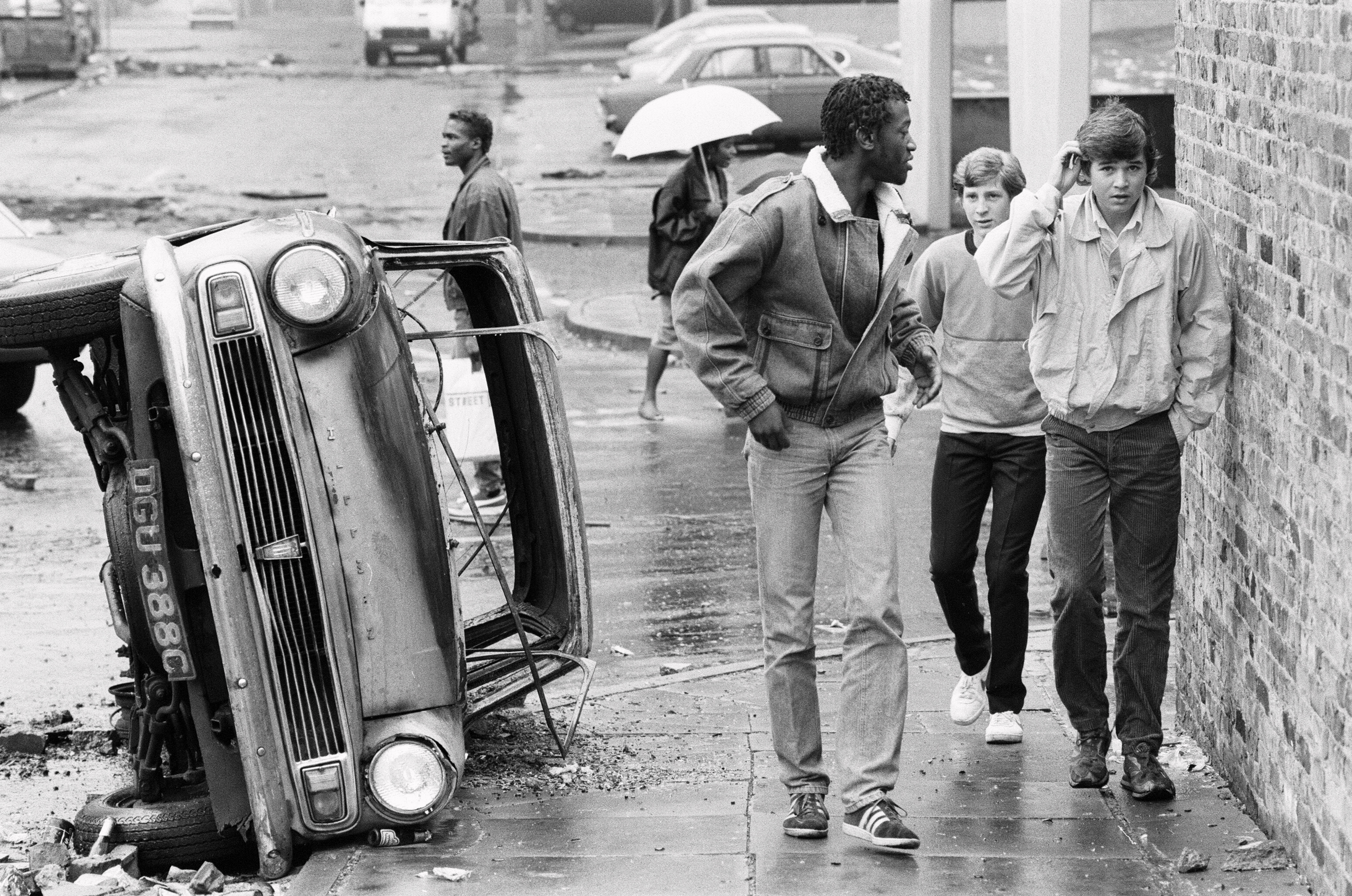 Aftermath of the riots which broke out in the Broadwater Farm estate in Tottenham, North London. The riot started the day after local resident Cynthia Jarrett died during a disturbance while police searched her home. Picture shows three youths walking past the wreckage of a burnt out car in Broadwater Farm, the day following the riot. 7th October 1985. (Photo by Andy Rosie/Mirrorpix/Getty Images)