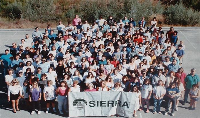 A company photo of Sierra, with dozens of people gathered in a parking lot behind a banner with the company logo in interlaced black and white and the legend SIERRA emblazoned on it.