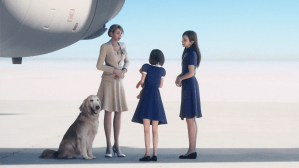 Screenshot from Ace Combat 7, a woman wearing an off-white dress and blazer stands next to a plane and a dog, with two younger looking women in blue dresses conversing with her.