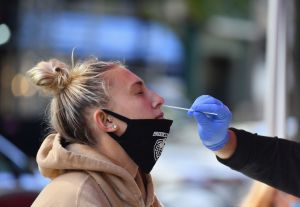 A woman in a brown hoodie with her blonde hair pulled back in a bun and a face mask lowered to her chin receives a nasal swab from a medical worker (not pictured except for their blue-gloved hand).