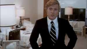 Screenshot from the movie "The Candidate," Bill McKay (played by a strawberry blond Robert Redford, wearing a dark blue suit) sits on the edge of a bed in a hotel room and asks his off-screen political advisor "what do we do next?"