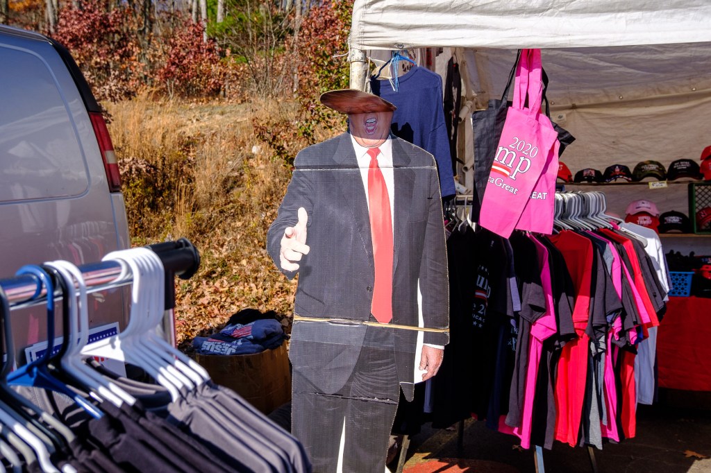 A cardboard cutout of Donald Trump stands at the opening to a merchandise booth full of Trump t-shirts and bags. The head of the cut-out is bent over from the wind.