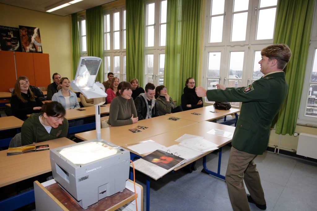 Ein Polizist gibt Präventions-Unterricht gegen Drogen an einer Schule, er steht vor der Klasse und bewegt die Hände wie ein Lenkrad