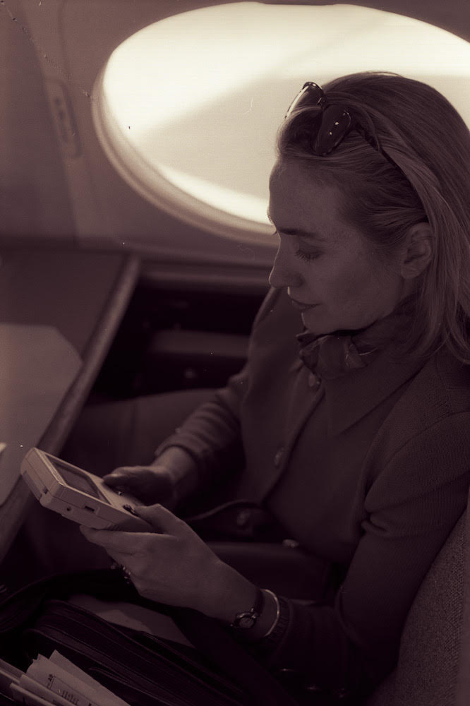 A sepia-toned black and white photo of Hillary Clinton in her First Lady years, playing on a Game Boy on Air Force One.