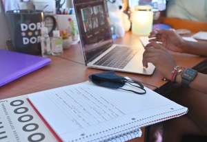 A student uses a laptop at a desk in their dorm room. A notebook and a cloth mask are on top of the desk