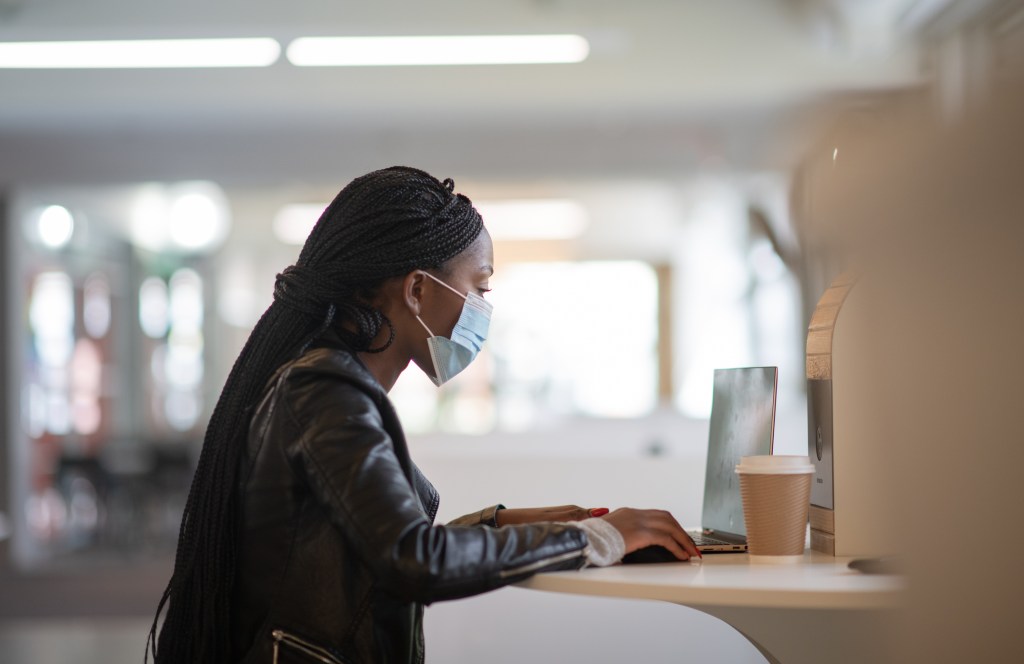 A Black woman sits at a desk in a well lit institutional office space and works on her laptop. She is wearing a protective mask.