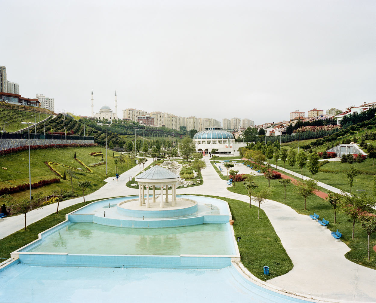 Mathias Depardon - Transanatolia. A garden with green medows, a fountain and pristine white and blue buildings.