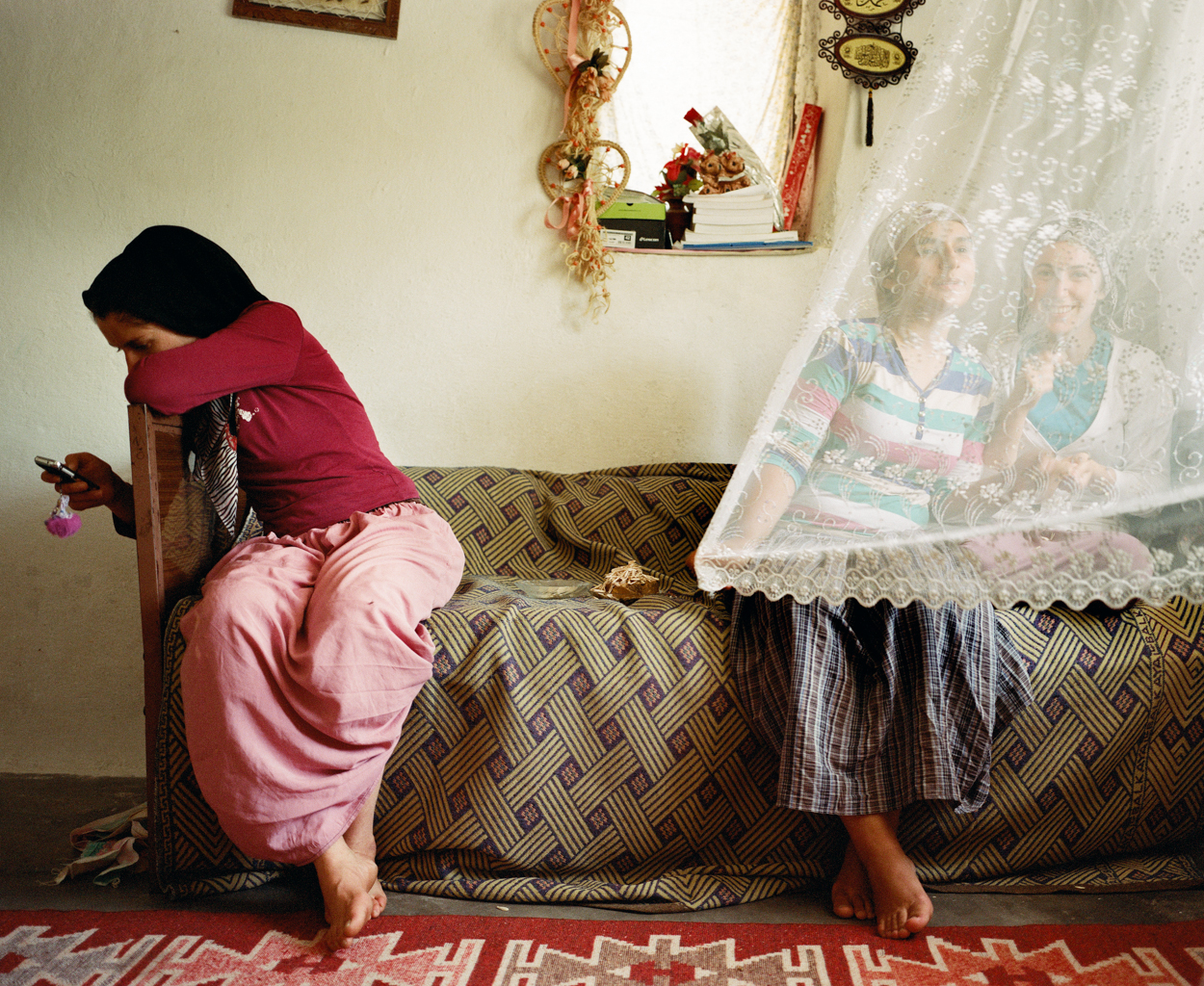 Mathias Depardon - Transanatolia. Three women sitting on a couch, one looking at her phone, the other two smiling at camera behind a lace curtain.