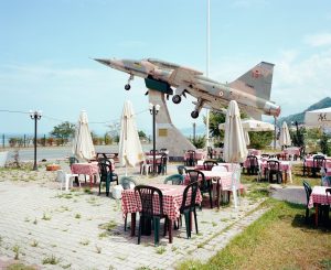 Die Terrasse eines Restaurants am Meer mit Tischen mit karierten Tischdecken und Plastikstühlen, dahinter an der Straße ist ein ausrangierter Düsenjäger aufgestellt.