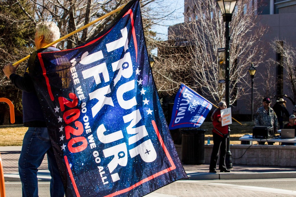 A man holds a flag reading :"Trump JFK JR. 2020."