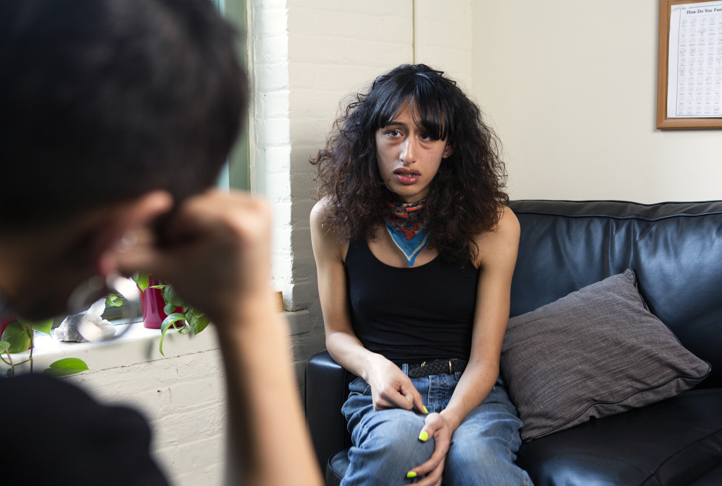 A feminine person wearing a tanktop and jeans sits on a black couch while talking to their therapist