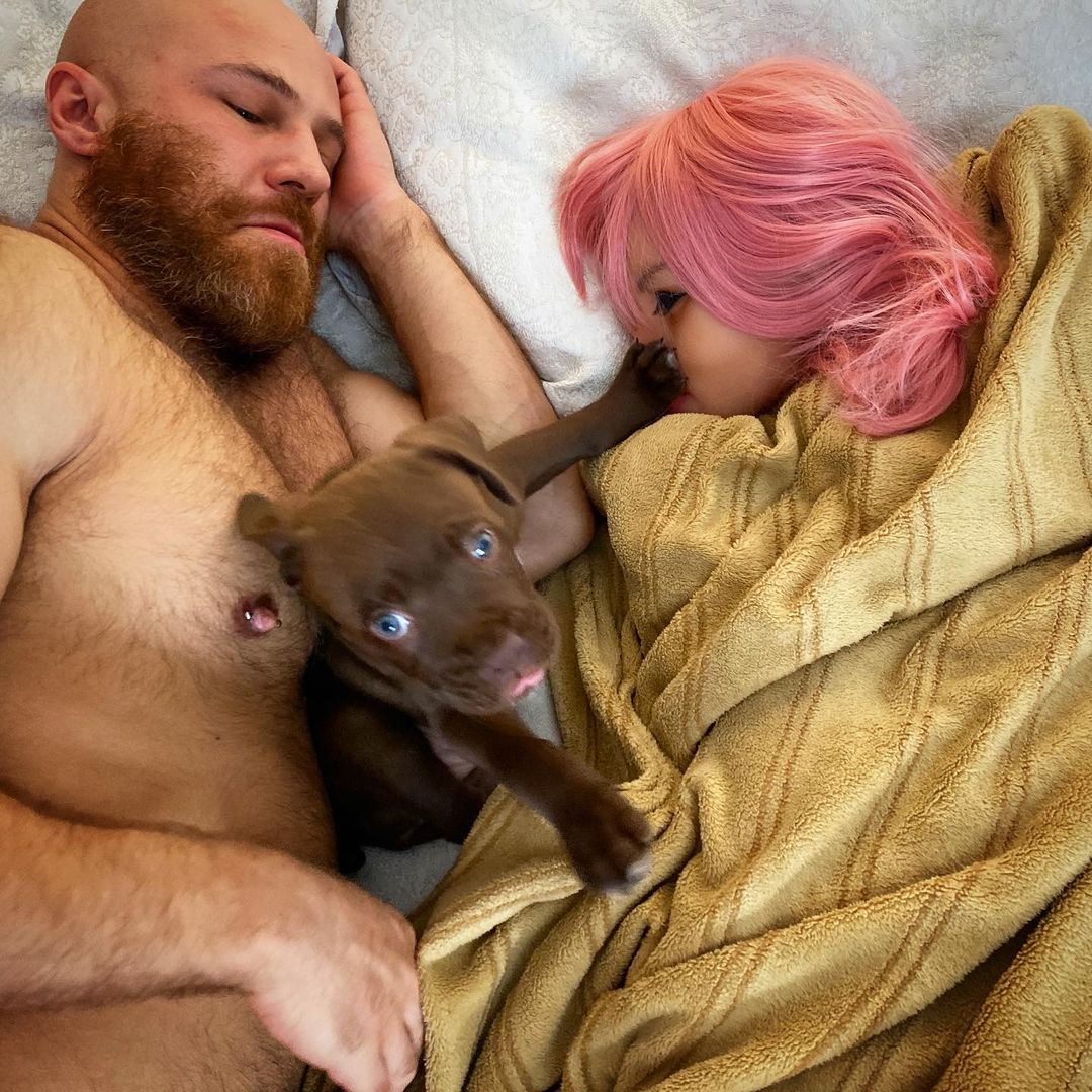 Ein muskulöse Mann mit nacktem Oberkörper im Bett mit einer Sexpuppe und einem Hund zwischen ihnen