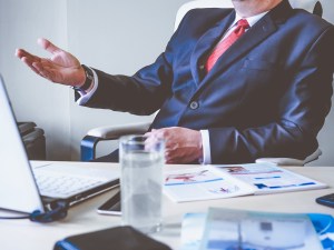 A man in a business suit and red tie extending his hand while sitting at a desk with a laptop