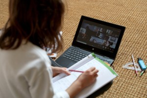 A students sits on the ground looking at a laptop while holding a notebook and pencil.