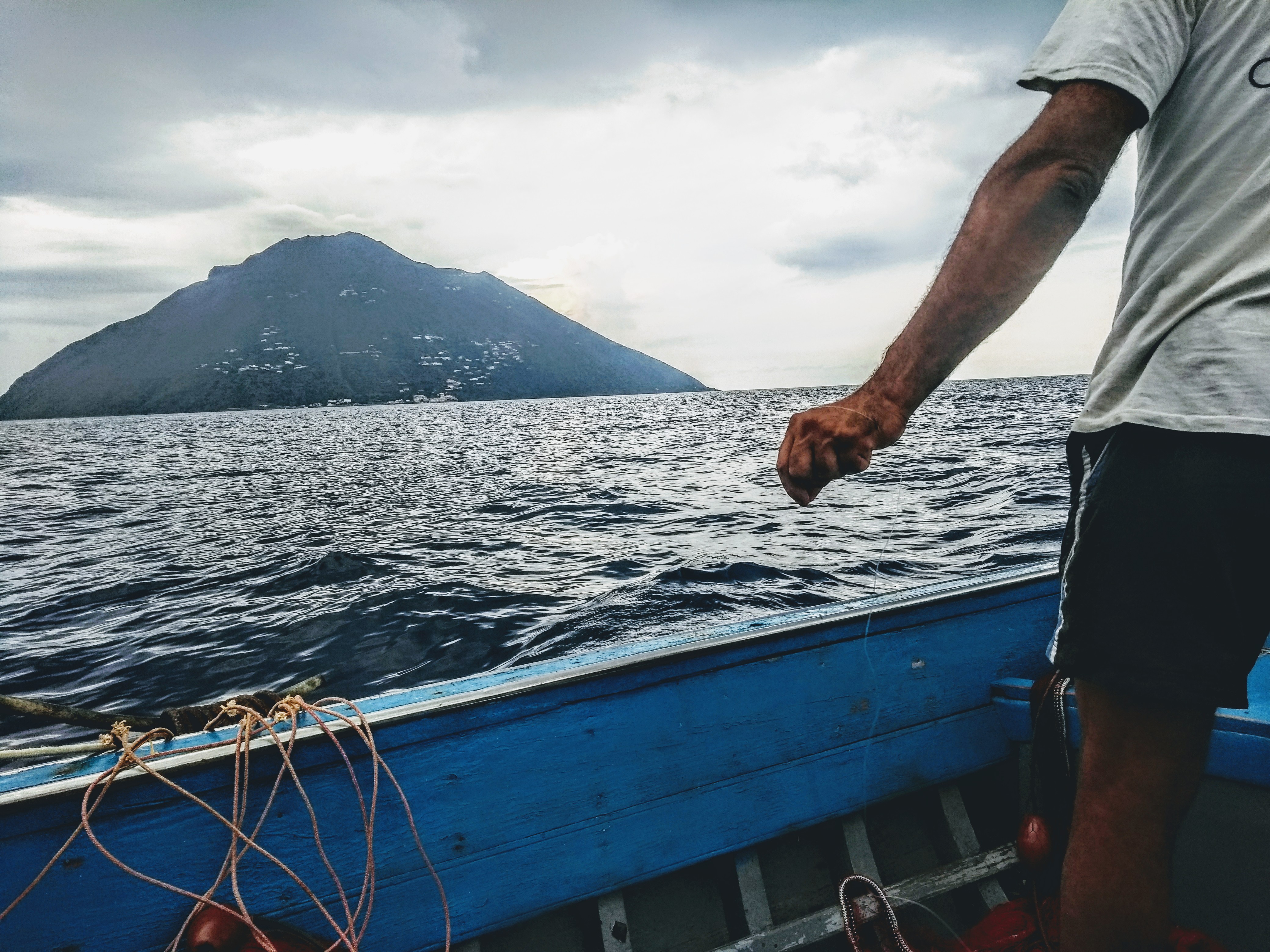 Alicudi island – Picture of a man with his back to us, throwing a fishing line into the water from his small blue wooden boat.