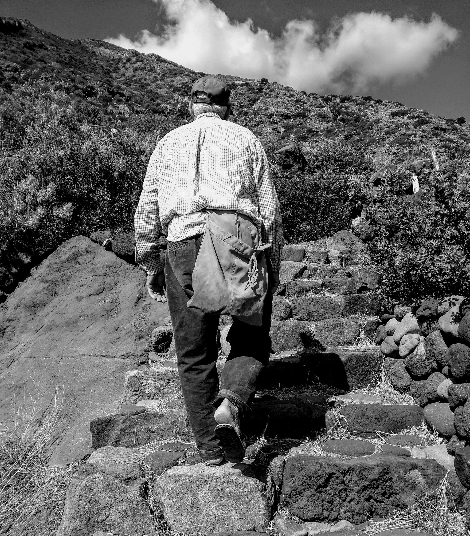 Alicudi island – black and white picture of an older man wearing sandles and a hat, climbing up some steep steps made out of stone.