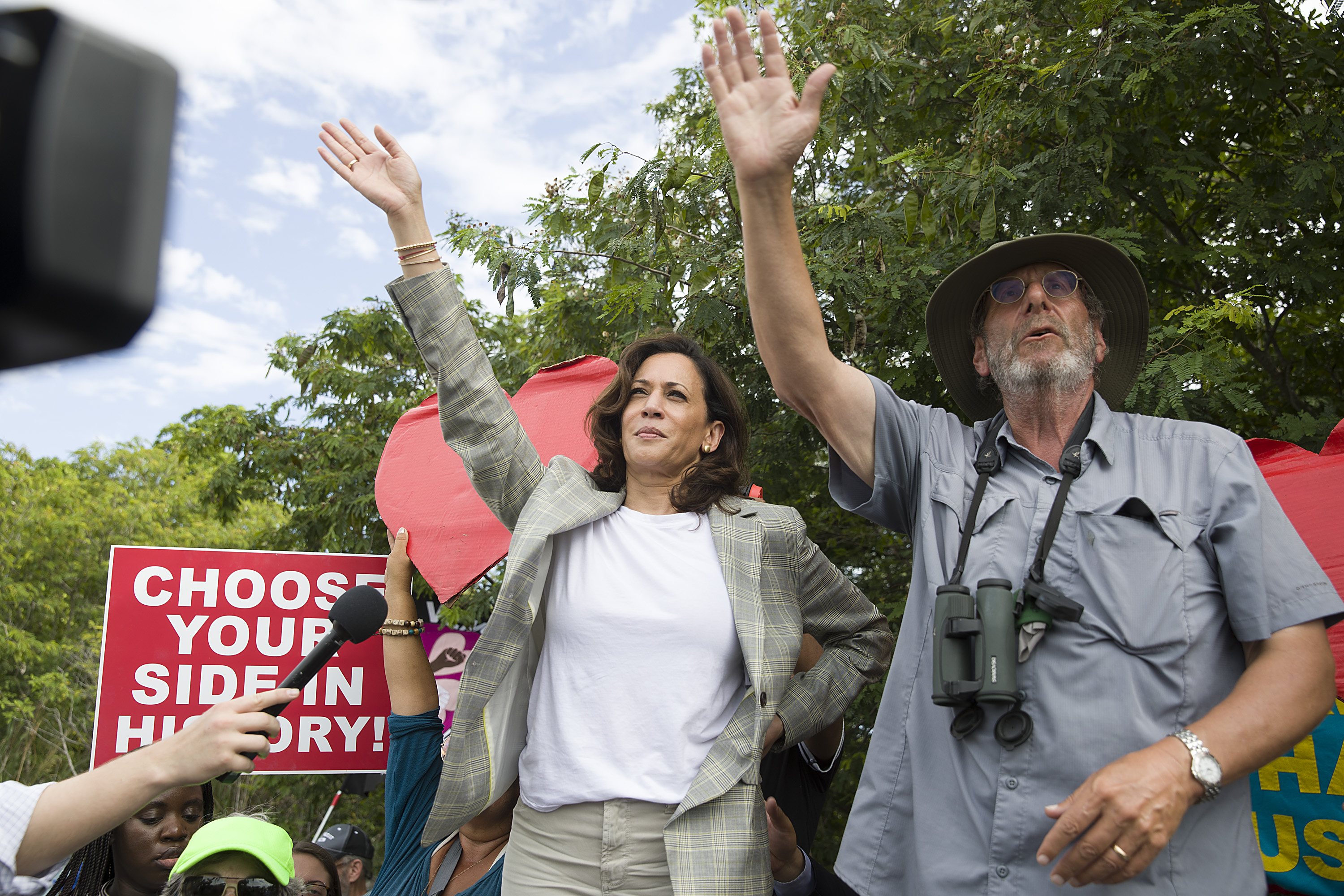 Kamala Harris waves as she stands on a ladder to look over a fence to see the detained migrant children at Homestead on June 28, 2019