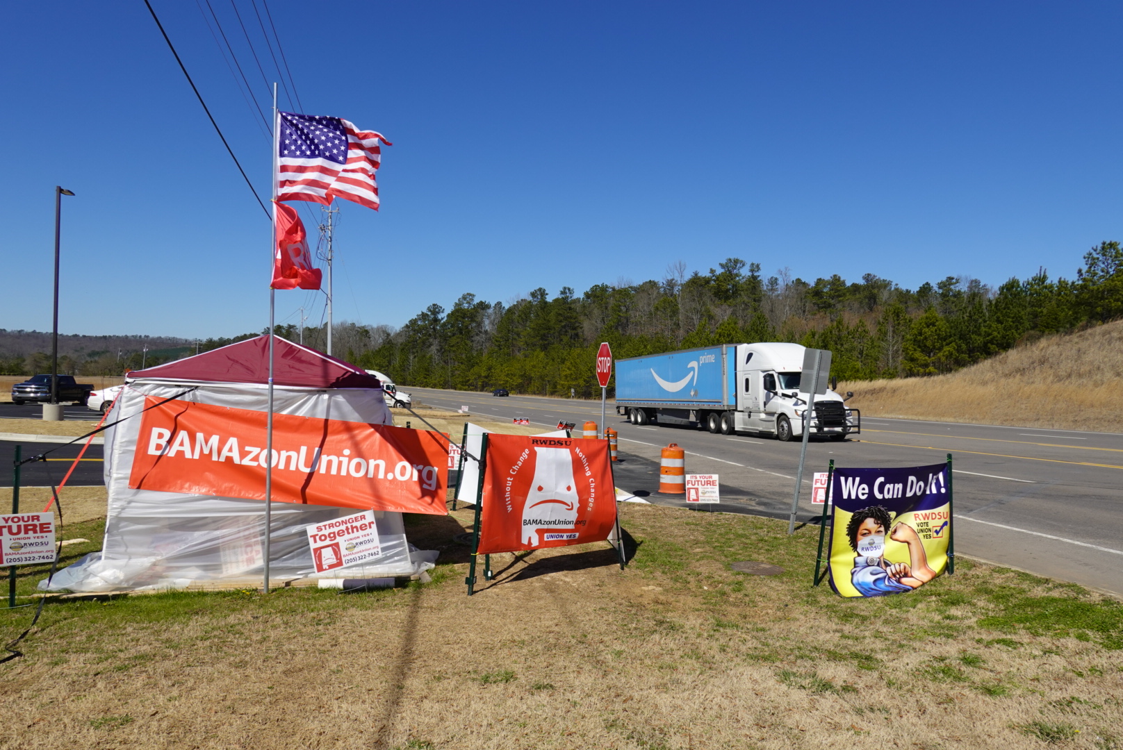 A red tent on the side of a road. There is a BAMAzonUnion.org poster, an American flag, and a We Can Do It! poster on the tent. An amazon truck drives by.