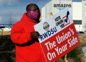 A Black man wearing a mask and sweatshirt holds a RWDSU VOTE YES sign in front of an A