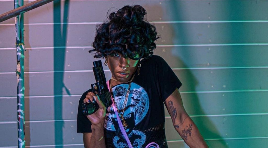 A Black woman wearing a black graphic t-shirt poses with a handgun in front of backdrop lit with pink and green lights.