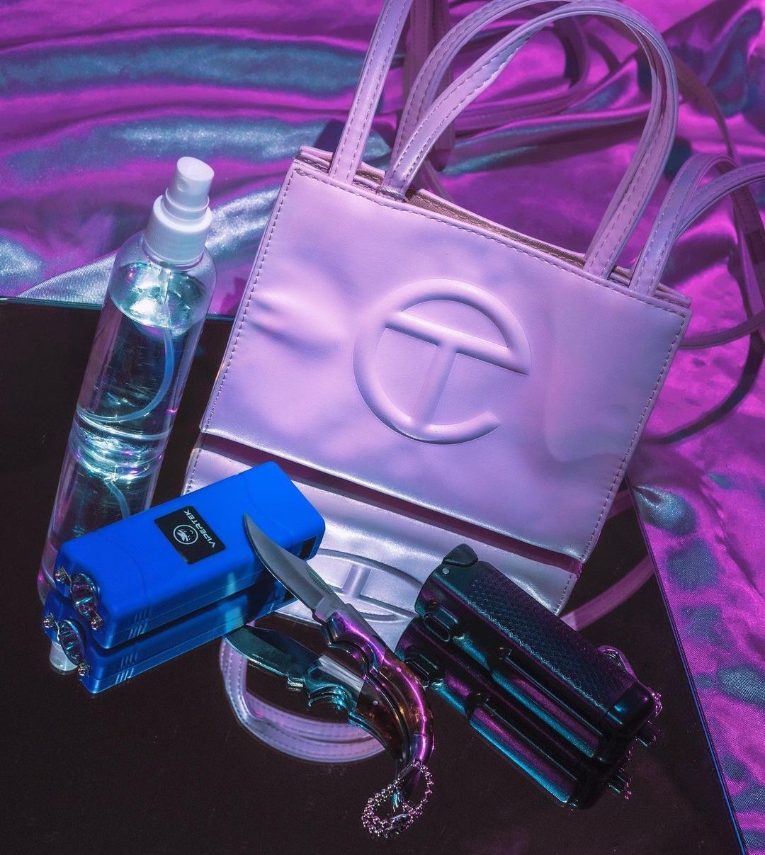 Self-defense items sit on a reflective table in front of a pink and blue background, including a bottle of pepper spray, a stun gun, a switchblade, and a white Telfar tote bag.