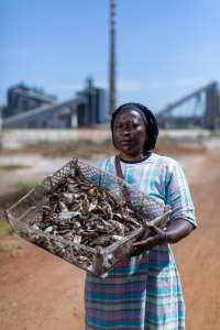 A woman stands holding fish in front of a coal-firedpower station