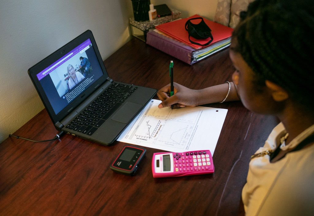 A girl wearing a yellow shirt sits at a brown wooden desk and writes on paper while attending a virtual class on her laptop