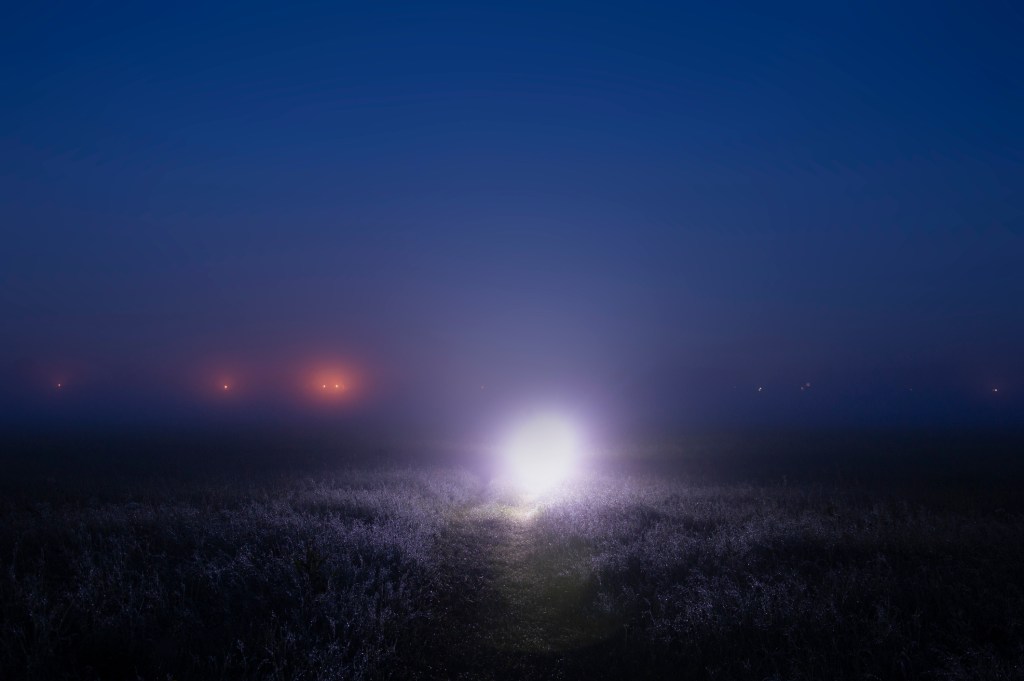 A mysterious bright light on a path on a misty night, with glowing lights on the horizon.