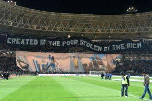 Black banner with white letters stretched across the Curva Nord of the Stade Olympique de Radès in Tunis. Bottom: another large banner with drawings.