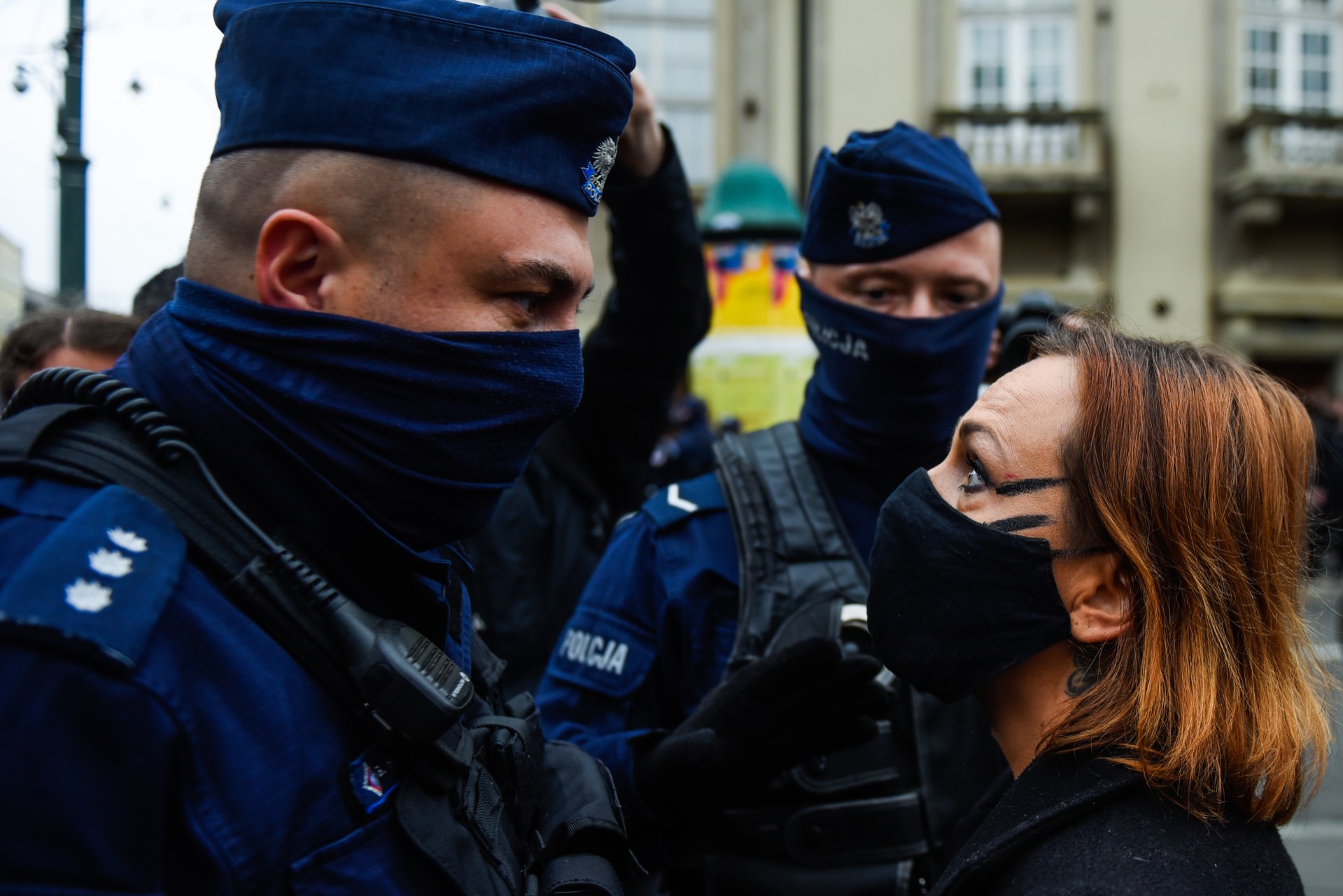 Zwei maskierte Polizeibeamte stehen einer Frau mit Gesichtsmaske gegenüber