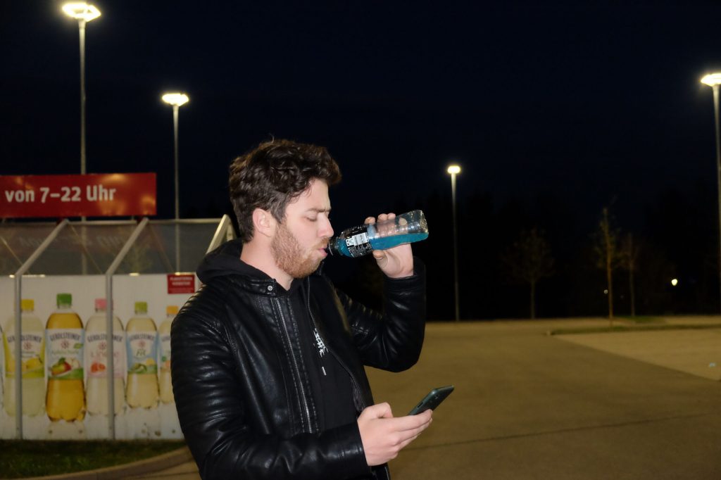 Junger Mann auf dunkler Straße trinkt Powerade und schaut aufs Handy