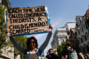 An anti-lockdown protester holds a sign that says, "Each time we're called conspiracy theorists, a child is mentally scarred. Tick tock..."