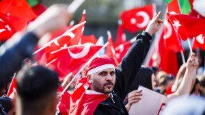 Ein Mann, der sich eine Türkeiflagge um den Kopf gebunden hat und umgeben ist von türkischen Flaggen macht das Handzeichen der grauen Wölfe, die in deutschland immer mehr zu einer Gefahr werden