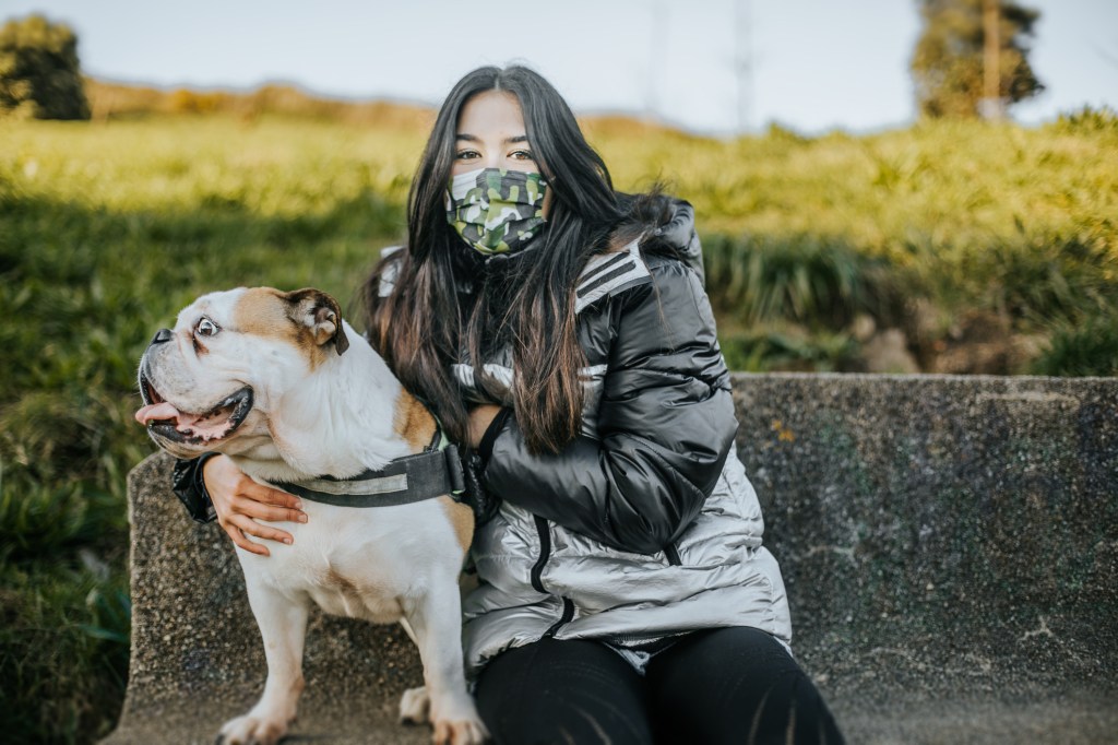 young woman sport clothes sitting in stone bench with dog besides she is wearing a protective mask in pandemic times