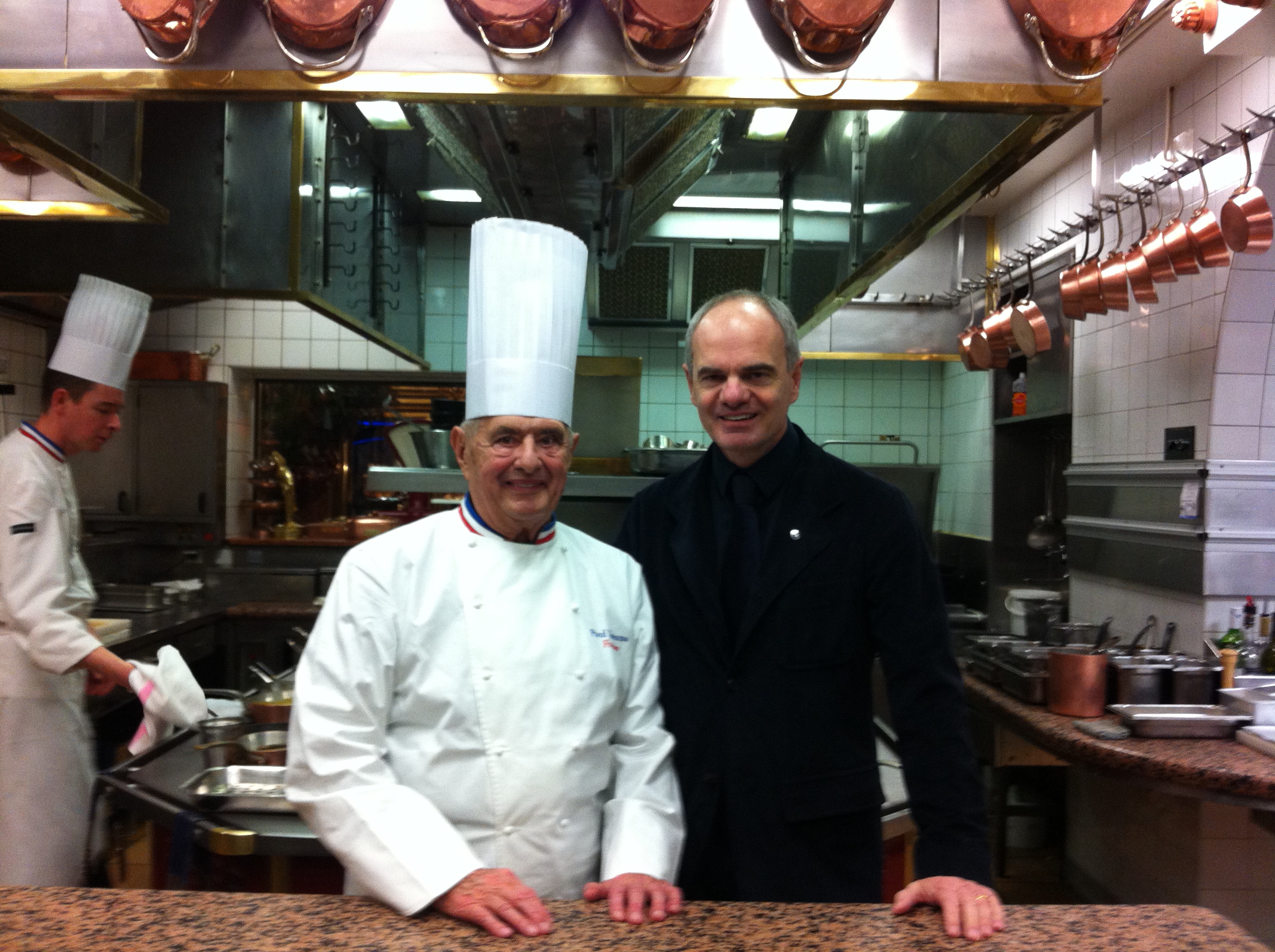 Enrico Derflingher – picture of a man in a white chef's uniform standing next to Derflingher (in black) in a large professional kitchen as a young cook is working in the background.