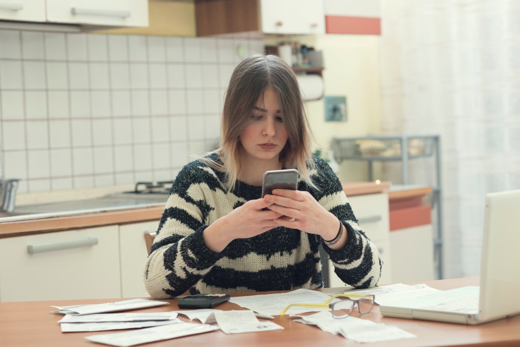 Young woman uses her phone while sitting at a table paying her bills