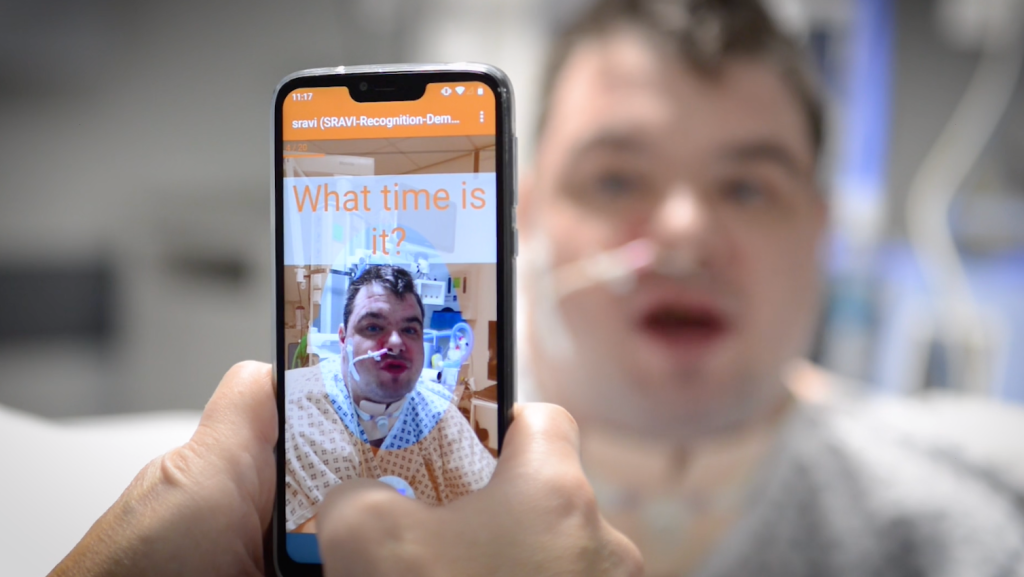 A person holds up a smartphone in front of a person with an oxygen tube in their nose. Text on the screen shows the phrase "what time is it?"