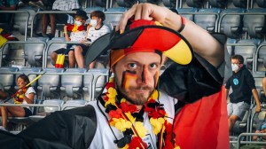 Vice-Autor Robert Hofmann in Deutschland-Merchandise gekleidet schaut traurig in die Kamera, hinter ihm trauernde Fans im Stadion. Es ist schön, dass die Deutschen jetzt traurig darüber sind, bei der EM verloren zu haben.