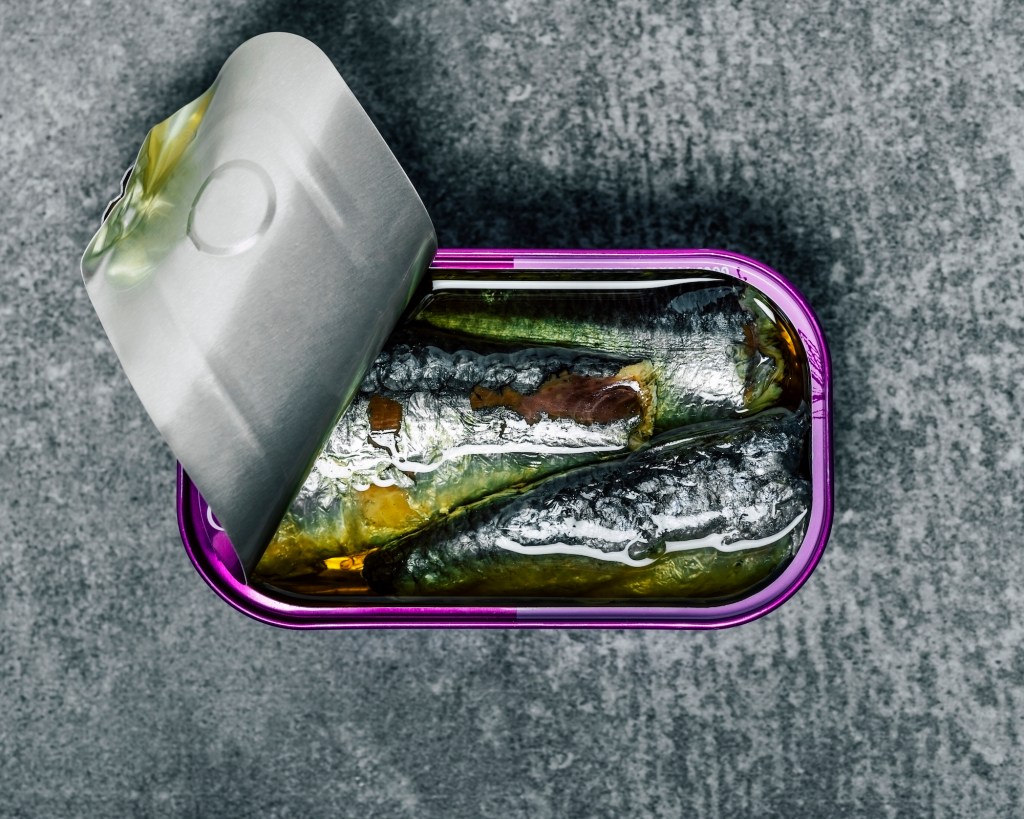 a purple can of sardines in front of a textured gray background; the tin is partially open, showing three fish inside