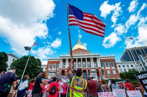 A man with a QAnon vest and US Flag joins hundreds of people of mixed political views, religions and cultures as they protest a mandate from the Massachusetts Governor requiring all children,age K-12, to receive an influenza (flu) vaccine/shot to attend s