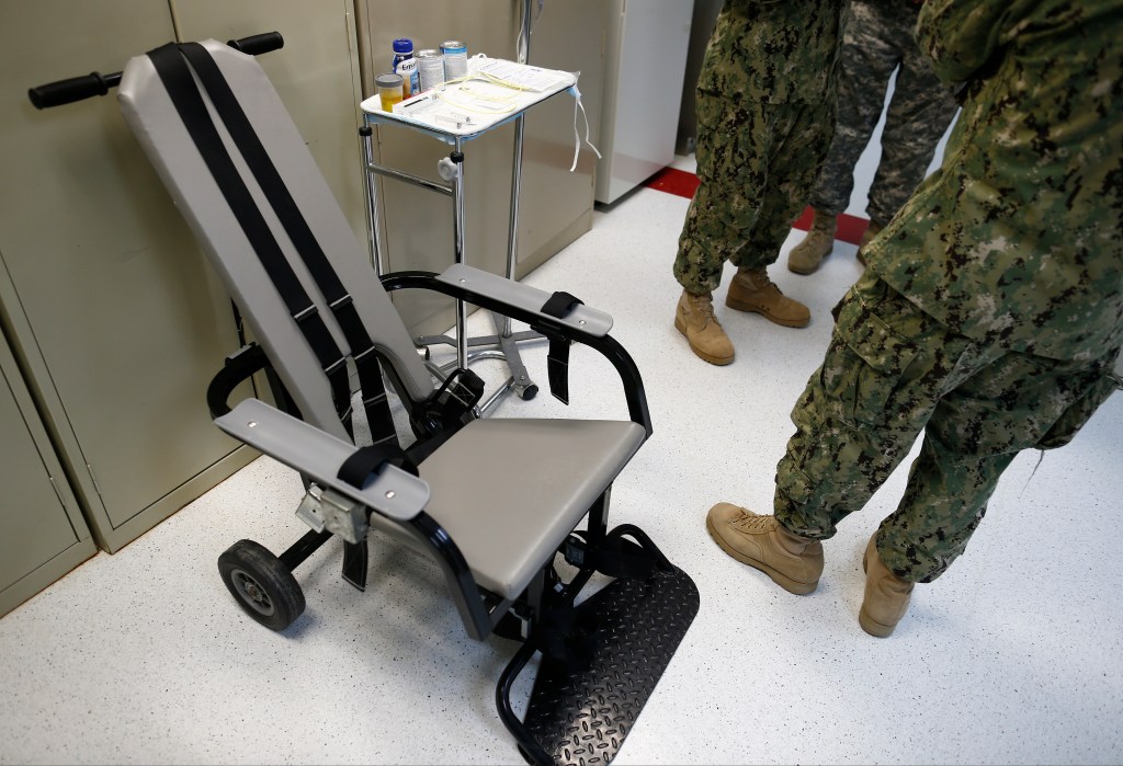 In this photo reviewed by the U.S. military, a U.S. Navy medical personel stand next to a chair with restraints, used for force-feeding.