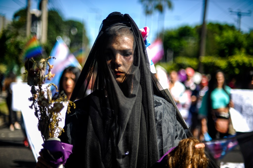 A trans woman wearing a black veil holds a wig and dead flowers while protesting violence in El Salvador.