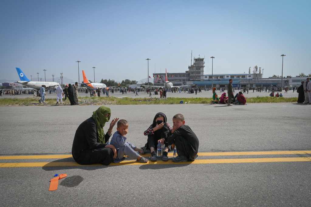 Afghan people sit along the tarmac as they wait to leave the Kabul airport in Kabul on August 16, 2021, after a stunningly swift end to Afghanistan's 20-year war, as thousands of people mobbed the city's airport trying to flee the group's feared hardline