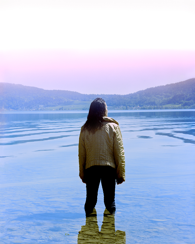 Mathias de Lattre, France, magic mushrooms - the author's mother, wearing a beige coat, stood in a shallow body of blue water, looking toward's a light pink sky.