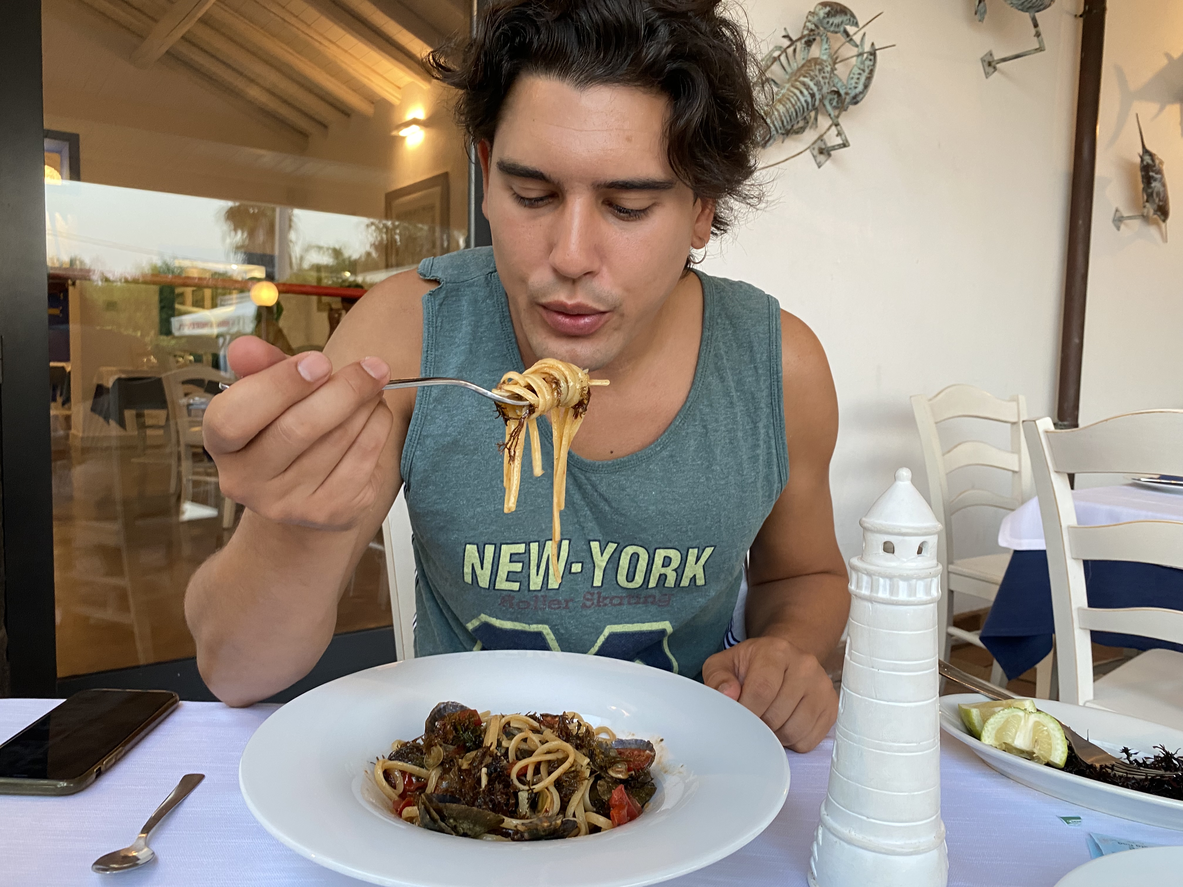 Mauru, Catania, Sicily – Man seated at a table decorated with a marine theme, about to eat a forkful of spaghetti with seaweed, various shellfish and cherry tomatoes.