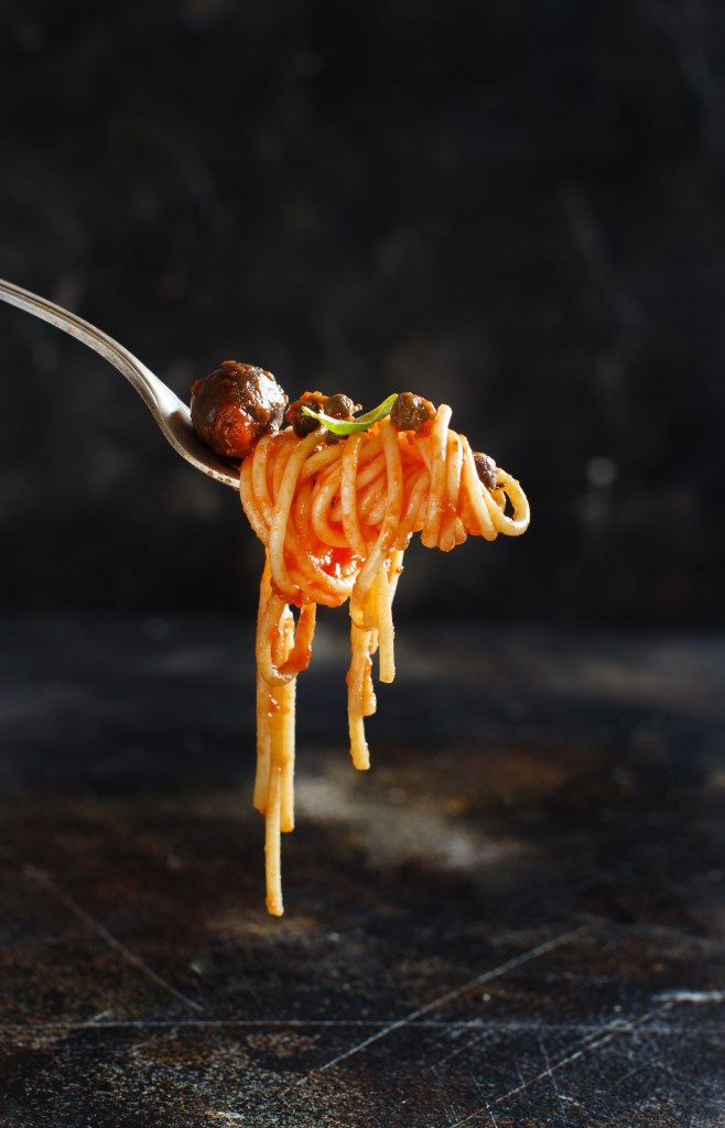 Pasta alla Puttanesca – fork holding up spaghetti in tomato sauce, a black olive and capers.