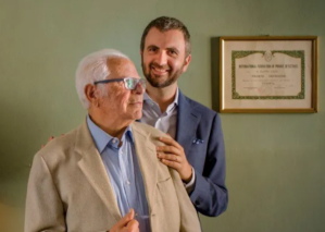 Mario and Francesco Caliò, private investigators, Italy - Bearded man in blue suit stands with hand on the shoulder of an older man wearing a white suit jacket, both posed in front of green wall.