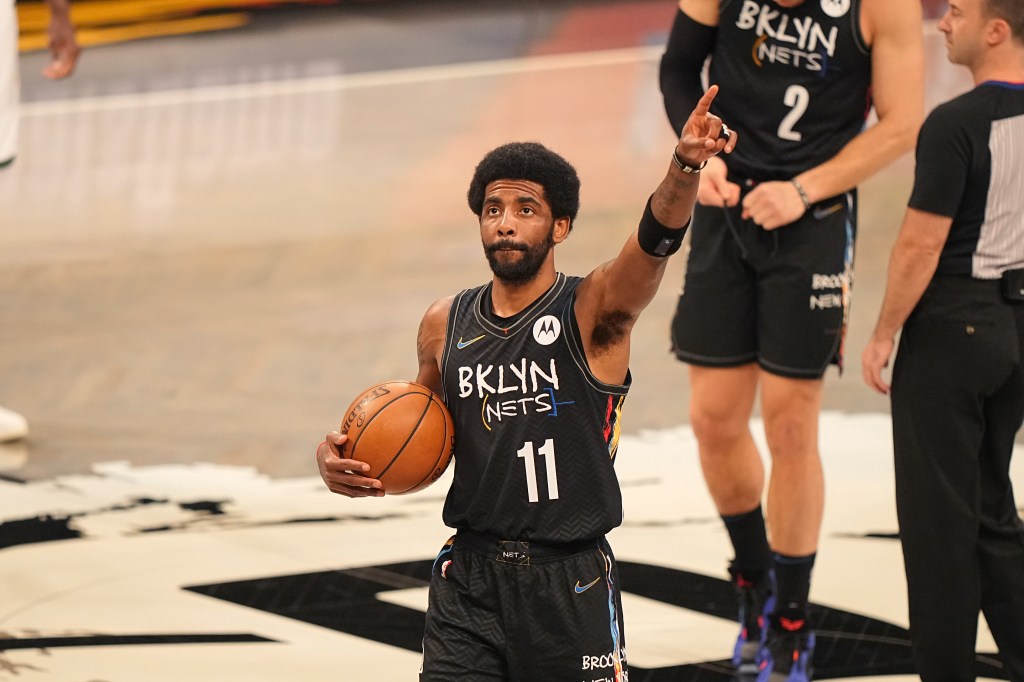 Kyrie Irving is shown on a basketball court, holding a basketball in one hand and raising the other arm, a finger pointed in the air.