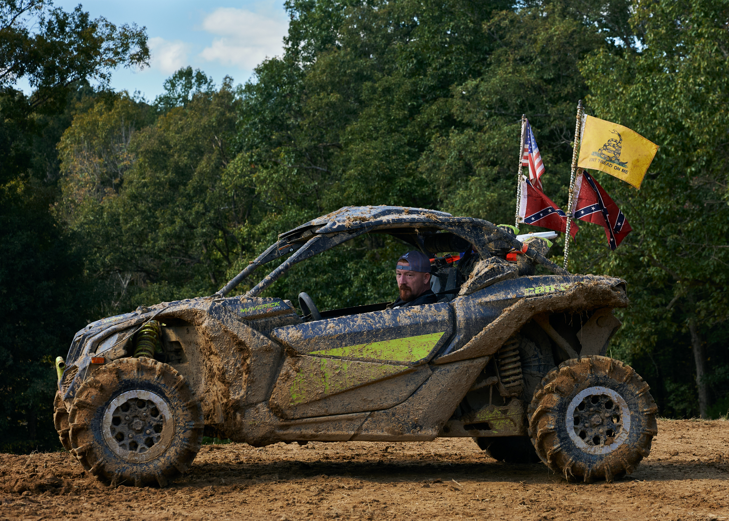 Ein Mann mit umgedrehter Baseball-Cap sitzt in einem mit Matsch zugekleisterten Offroad-Buggy, auf dem mehrere Flaggen befestigt sind