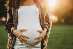 A pregnant person in a white top is seen holding their stomach. Their face is not visible. A second person stands behind them and clasps their hips. There's a hazy sunset in the background.